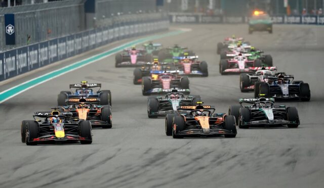 Group of colorful Formula 1 cars racing side by side on a track, near the start of the race with barriers along the left.