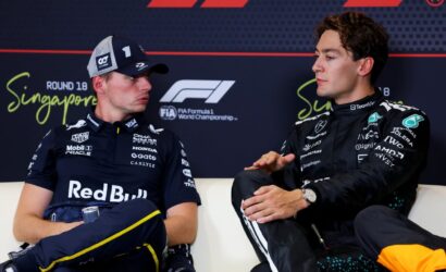 Two Formula 1 drivers sit on a white couch and talk during a press session in Singapore, wearing team racing suits and caps with sponsor logos, with a FIA F1 backdrop behind them (Singapore Grand Prix).