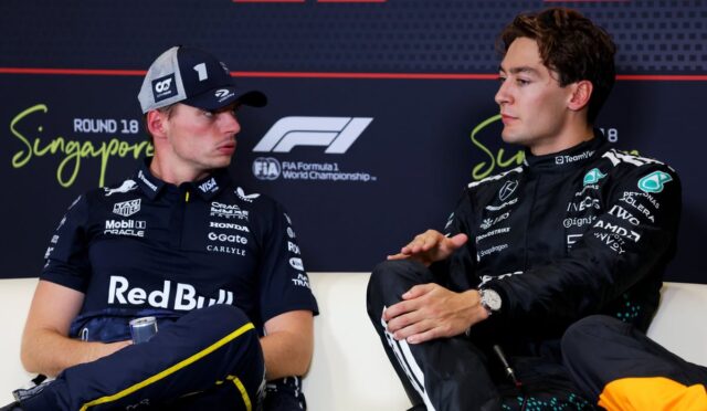 Two Formula 1 drivers sit on a white couch and talk during a press session in Singapore, wearing team racing suits and caps with sponsor logos, with a FIA F1 backdrop behind them (Singapore Grand Prix).