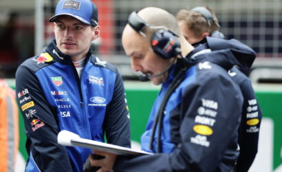 Two Formula 1 engineers in blue racing jackets study a clipboard during a pit stop, one wearing a cap and the other with a headset.