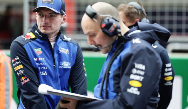 Two Formula 1 engineers in blue racing jackets study a clipboard during a pit stop, one wearing a cap and the other with a headset.