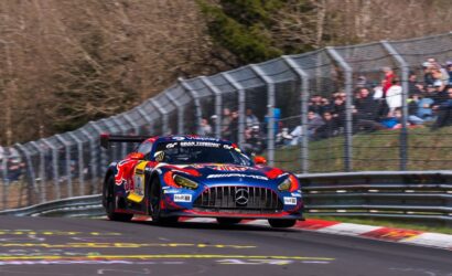 Front view of a blue and red Mercedes-AMG GT3 race car taking a high-speed corner on a race track, with spectators behind a safety fence.