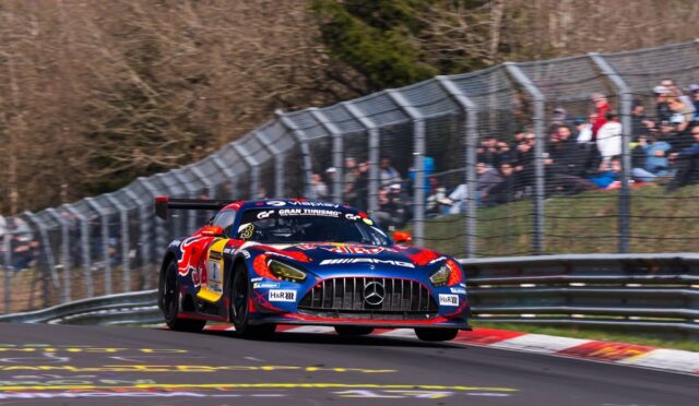 Front view of a blue and red Mercedes-AMG GT3 race car taking a high-speed corner on a race track, with spectators behind a safety fence.