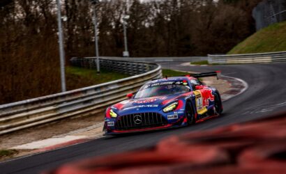 Red Bull–sponsored Mercedes race car rounding a curving track, headlights on, barrier rails on the side of the road.