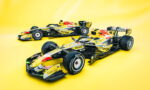 Two Red Bull Racing Formula 1 cars on a solid bright yellow backdrop, angled as if ready to race (yellow and black livery).