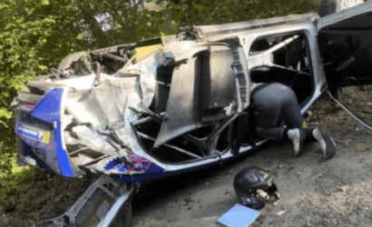 A blue and white van lies on its side after a severe crash, with a rescuer kneeling inside the open door area amid debris, helmet and gear on the ground nearby.
