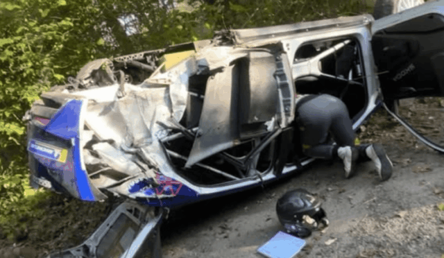 A blue and white van lies on its side after a severe crash, with a rescuer kneeling inside the open door area amid debris, helmet and gear on the ground nearby.