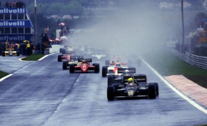 Leading black-and-gold Formula 1 car on a rain-soaked track, No.12, with spray from tires and a pack of cars behind