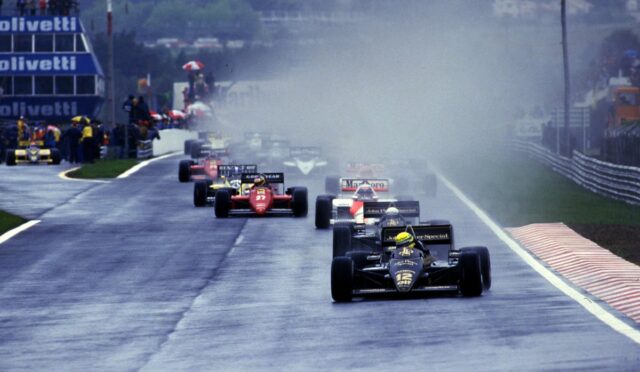 Leading black-and-gold Formula 1 car on a rain-soaked track, No.12, with spray from tires and a pack of cars behind
