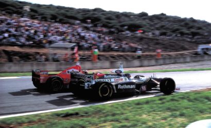Open-wheel race car with Rothmans and Castrol sponsorship speeding on a track, crowd in stands.