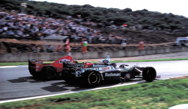 Open-wheel race car with Rothmans and Castrol sponsorship speeding on a track, crowd in stands.