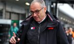 Man in a black jacket with F1 logo and Tommy Hilfiger badge, wearing glasses, pointing with his index finger in an outdoor motorsport setting.