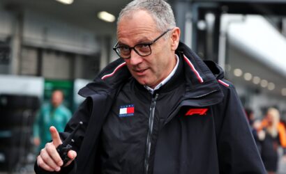 Man in a black jacket with F1 logo and Tommy Hilfiger badge, wearing glasses, pointing with his index finger in an outdoor motorsport setting.