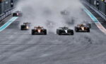 Formula 1 cars sprint from the start on a damp track, kicking up a large spray/smoke cloud as orange-and-black cars lead the pack.