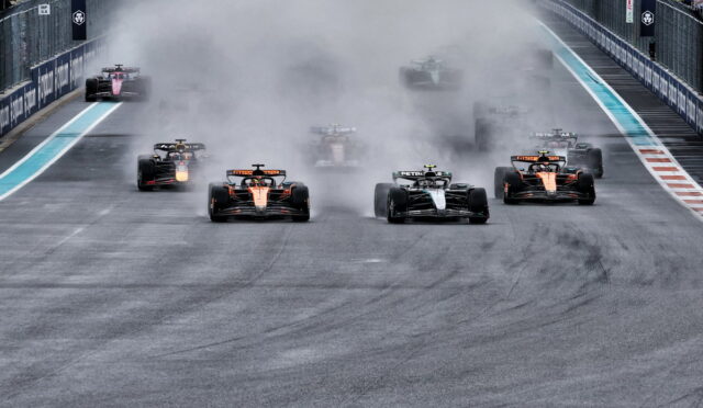 Formula 1 cars sprint from the start on a damp track, kicking up a large spray/smoke cloud as orange-and-black cars lead the pack.