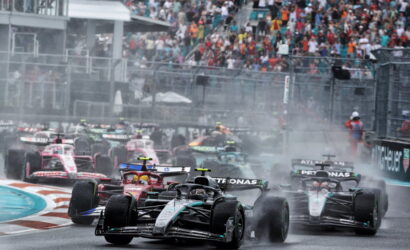 Multiple Formula 1 cars race closely on a wet track, spraying water as they speed toward a turn with a crowded grandstand in the background.