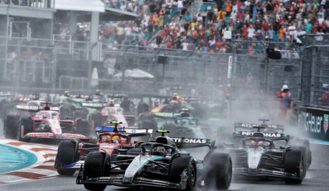 Multiple Formula 1 cars race closely on a wet track, spraying water as they speed toward a turn with a crowded grandstand in the background.