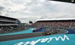 View of a Formula 1 race on a blue track with multiple cars entering a curve under a cloudy sky, grandstands full of spectators in the background.