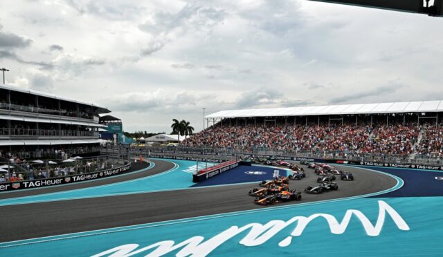 View of a Formula 1 race on a blue track with multiple cars entering a curve under a cloudy sky, grandstands full of spectators in the background.