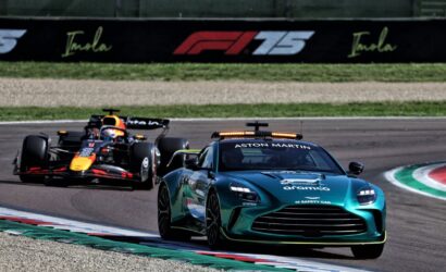 A teal Aston Martin safety car leads a Formula 1 race, with a Red Bull Racing car close behind on a curving track.