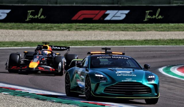 A teal Aston Martin safety car leads a Formula 1 race, with a Red Bull Racing car close behind on a curving track.