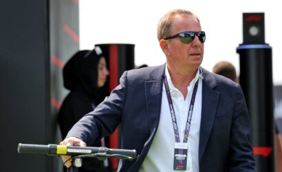 Man in a navy blazer and sunglasses stands outdoors, gripping the handlebar of an electric scooter and wearing a lanyard badge at an event.