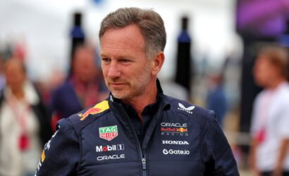 Man in a navy Red Bull Racing jacket with sponsor logos standing outdoors, looking to the side.