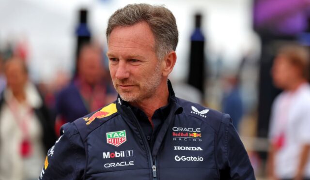 Man in a navy Red Bull Racing jacket with sponsor logos standing outdoors, looking to the side.