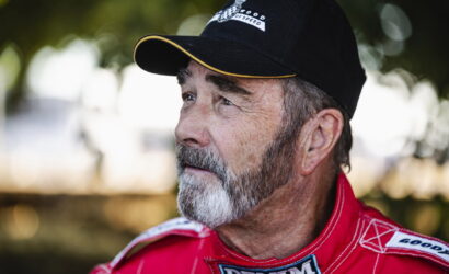 Older man with a gray beard in a red racing suit, outdoors, looking to the right.