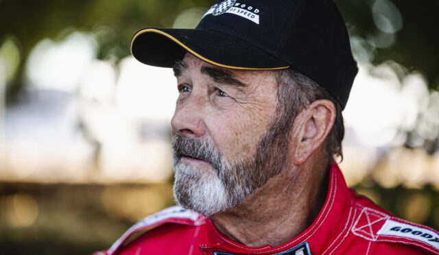 Older man with a gray beard in a red racing suit, outdoors, looking to the right.