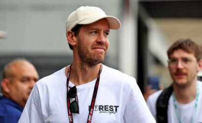 Man in a white cap and t-shirt with a lanyard, looking to the right at an outdoor event, sunglasses hanging from his shirt.