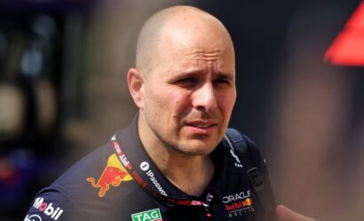 Close-up portrait of a bald man in a dark racing suit with sponsor logos, looking focused and tense.