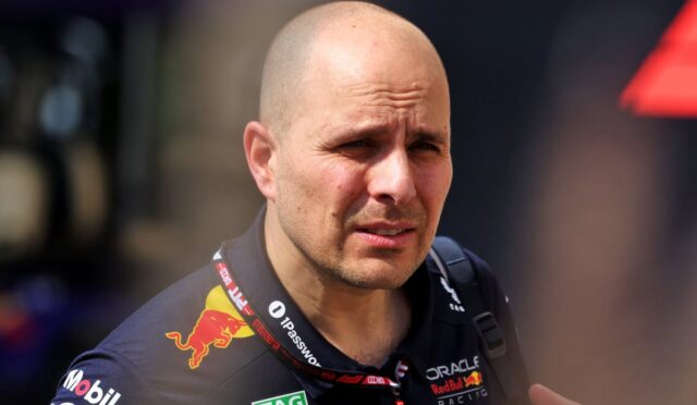 Close-up portrait of a bald man in a dark racing suit with sponsor logos, looking focused and tense.