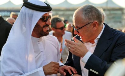 Two men talk and laugh at an outdoor event; one wears a white kandura and sunglasses, the other a dark suit and glasses.
