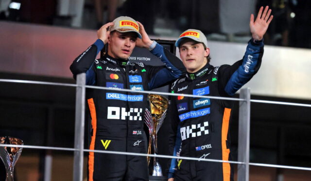 Two race car drivers on a podium in dark team suits with Pirelli caps, celebrating with a large trophy nearby and one waving hands up.