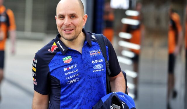 Man in a blue Red Bull racing polo smiling, carrying a helmet and backpack, in an outdoor area.