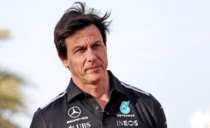 Man wearing a black Mercedes team polo, outdoors, with wind in his hair, looking thoughtful.