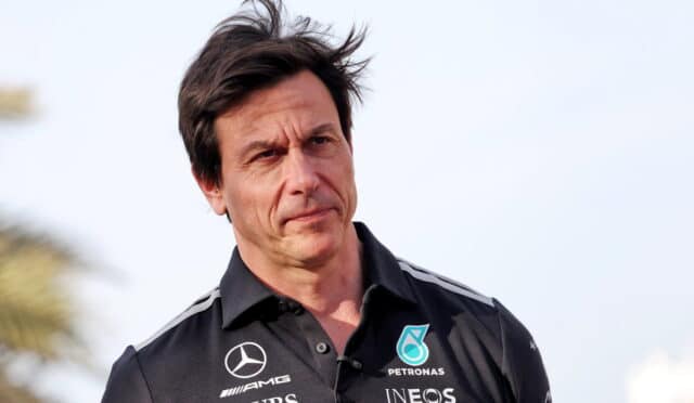 Man wearing a black Mercedes team polo, outdoors, with wind in his hair, looking thoughtful.