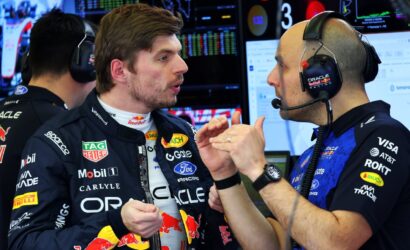 Two race car drivers in team uniforms wearing headsets share a fist bump in a busy pit garage filled with monitors and data screens behind them.