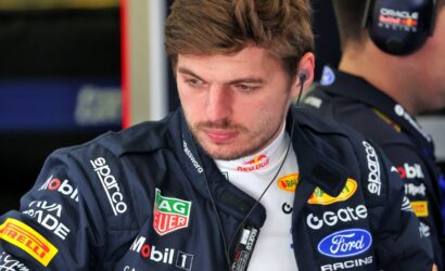 Race car driver in a sponsor-covered jumpsuit with headset, looking down.