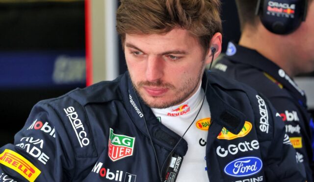 Race car driver in a sponsor-covered jumpsuit with headset, looking down.