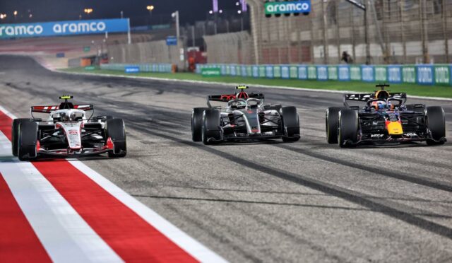 Three Formula 1 cars race side by side on a nighttime track, red and white curbs in the foreground and Aramco banners in the background.