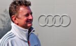 Smiling man in a white zip-up jacket with gray shoulder stripes, standing in front of a large Audi emblem.