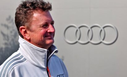 Smiling man in a white zip-up jacket with gray shoulder stripes, standing in front of a large Audi emblem.