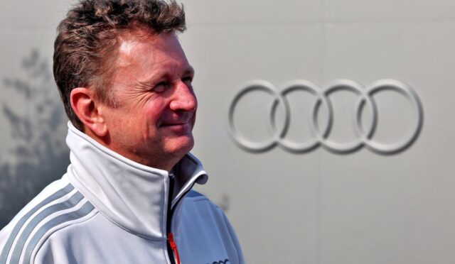 Smiling man in a white zip-up jacket with gray shoulder stripes, standing in front of a large Audi emblem.