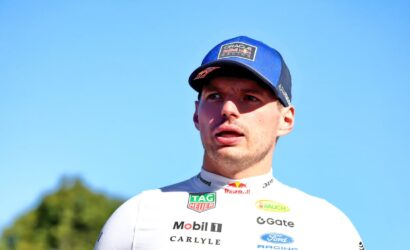 Portrait of a male race car driver in a white sponsor jersey and blue cap, looking to the side against a clear blue sky.