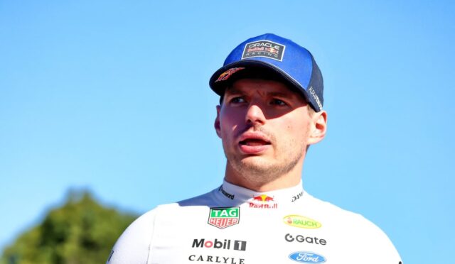 Portrait of a male race car driver in a white sponsor jersey and blue cap, looking to the side against a clear blue sky.