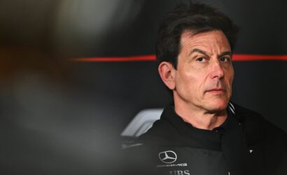 Portrait of a man in a black Mercedes‑AMG racing suit, looking to the side in a dim pit area.