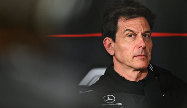 Portrait of a man in a black Mercedes‑AMG racing suit, looking to the side in a dim pit area.