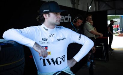 Race car driver in a white branded suit and black cap leans against a tire in a pit area, looking to the side.